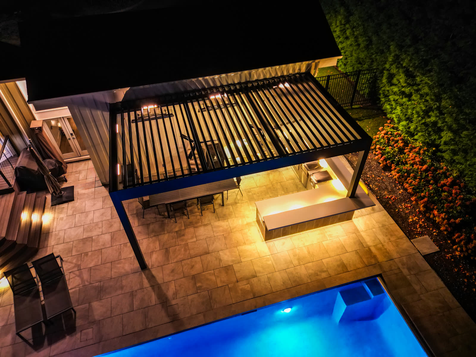 Nighttime aerial of lit louvered pergola beside glowing blue pool
